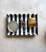 Siyah beyaz çizgili tepsi üzerinde saat, saat kutusu ve altın saat sergilenerek modern tasarım öğeleri ön plana çıkarılıyor.