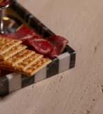 . Siyah beyaz çizgili modern mermer tepside peynir, üzüm, kraker ve meyveler yer alıyor.