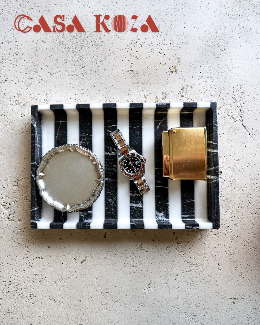 Siyah beyaz çizgili mermer tray üzerinde bir saat, saat kutusu ve ayna bulunuyor.